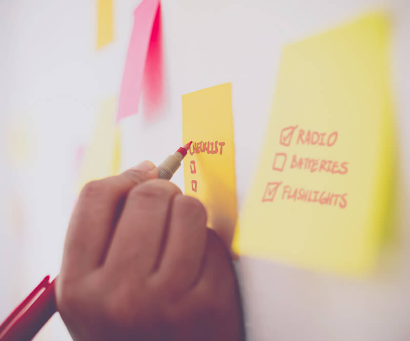A hand holding a red marker is checking off items like radio, batteries, and flashlights on a yellow sticky note labeled CHECKLIST. Other colorful notes—and a hurricane checklist for windows and doors—are visible on the wall.