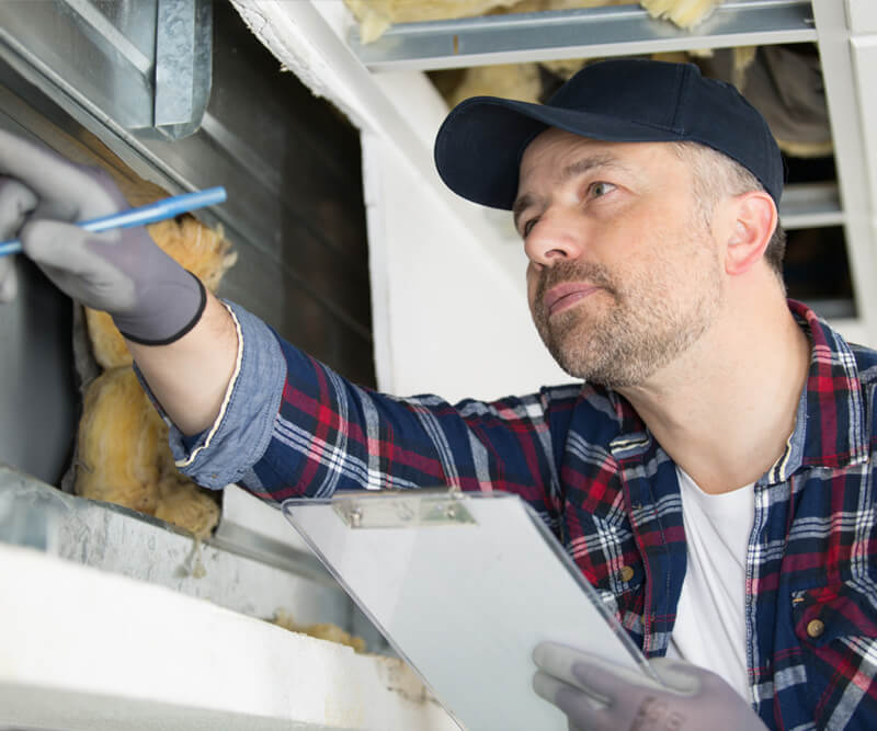 A man wearing a cap, flannel shirt, and gloves inspects an air duct with a clipboard and pen, possibly conducting a maintenance check as part of the My Safe Florida Home grants program.