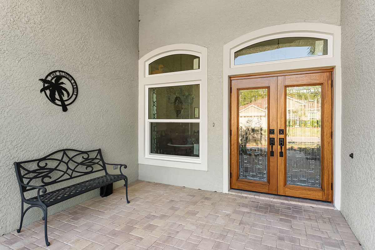 A covered front porch in Trinity,FL features a black metal bench, decorative wall art, and double wooden entry doors with glass panels—an inviting entry framed by a light-colored textured wall and a large window.