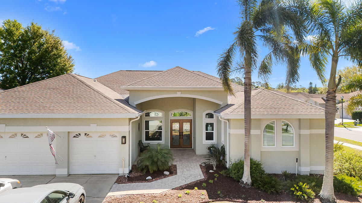 Single-story beige house in Trinity, FL with replacement windows and doors, a three-car garage, arched entryway, palm trees, and a manicured front yard under a clear blue Florida sky.