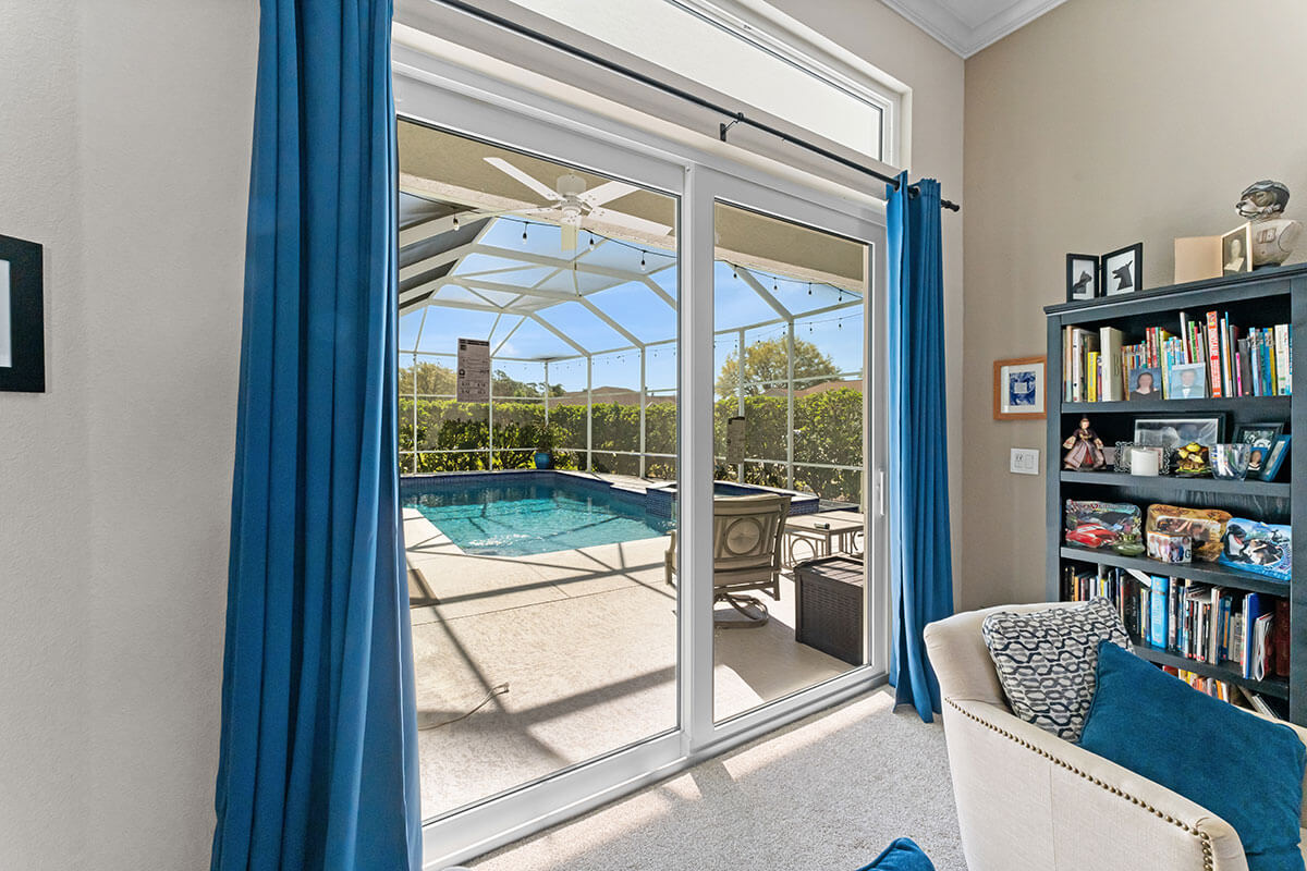 A bright room with a bookshelf, armchair, and blue curtains features sliding patio doors Florida-style, opening to a screened-in backyard pool with lounge chairs and lush greenery visible outside.