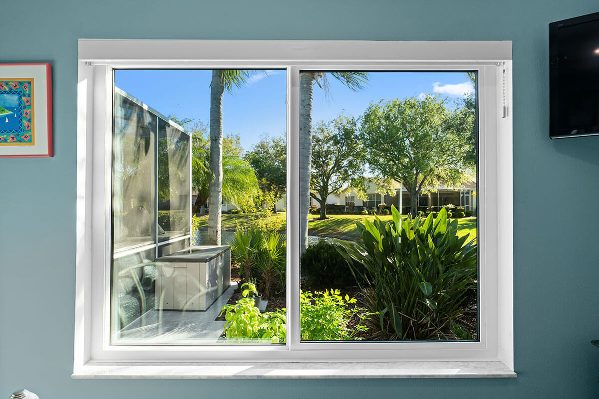 A large slider window with a white frame reveals a sunny Florida garden filled with lush green plants, palm trees, and a walkway outside. Part of the wall and a TV can be seen inside the room.