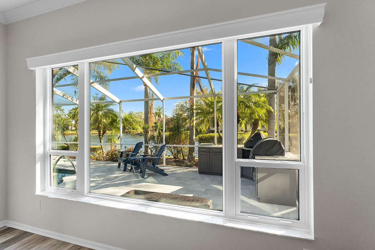 Large white-framed bay window looking out onto a sunny Florida patio with Adirondack chairs, a grill, and views of palm trees and a lake under a blue sky.