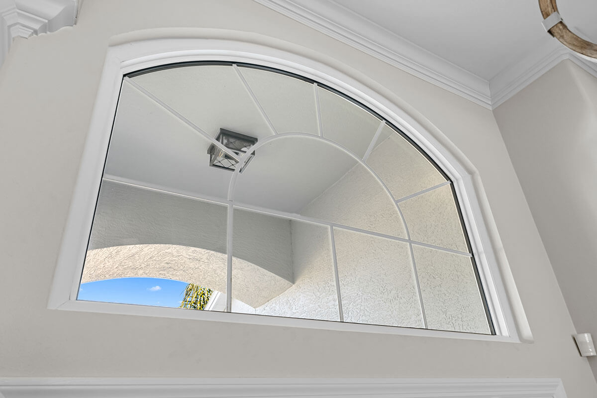 A large, arched, shaped window with grilles and white trim lets in natural light, reflecting an outdoor arch and a small section of blue sky. The wall is painted light gray, with crown molding framing the top edge.