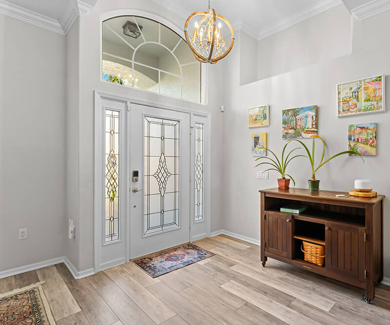 The spacious home entryway features a glass-paneled front door that welcomes you into a bright space adorned with a decorative chandelier. A wooden cabinet, embellished with potted plants and artwork, rests on light wood flooring, while a colorful rug adds charm.